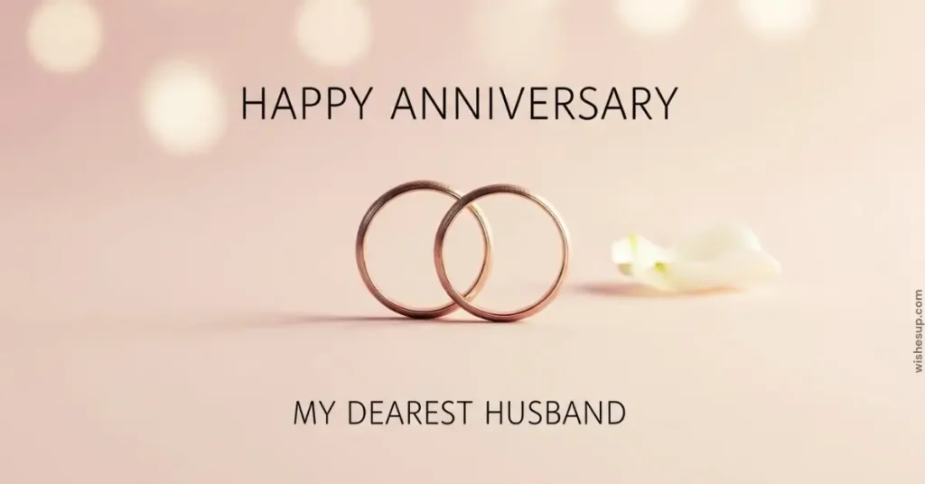 Two interlocking rose gold wedding bands rest on a soft, blush-pink surface beside a delicate white petal. Bathed in warm, diffused bokeh lighting, the scene features elegant black text that reads "HAPPY ANNIVERSARY MY DEAREST HUSBAND.