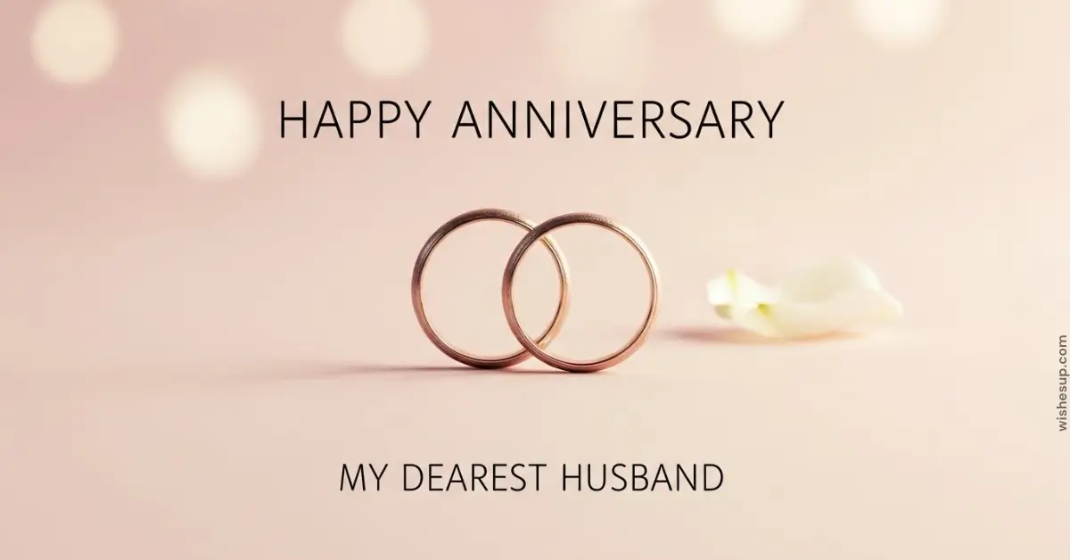 Two interlocking rose gold wedding bands rest on a soft, blush-pink surface beside a delicate white petal. Bathed in warm, diffused bokeh lighting, the scene features elegant black text that reads "HAPPY ANNIVERSARY MY DEAREST HUSBAND.