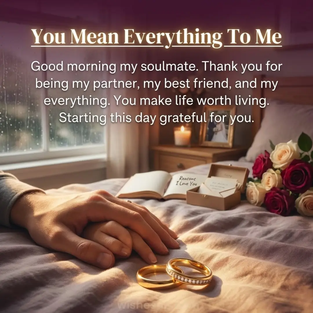 Intertwined hands rest on soft grey bedding beside two gleaming gold wedding rings, illuminated by warm morning sunlight. A bouquet of red and cream roses sits nearby, set against a rainy window and a glowing bedside candle.